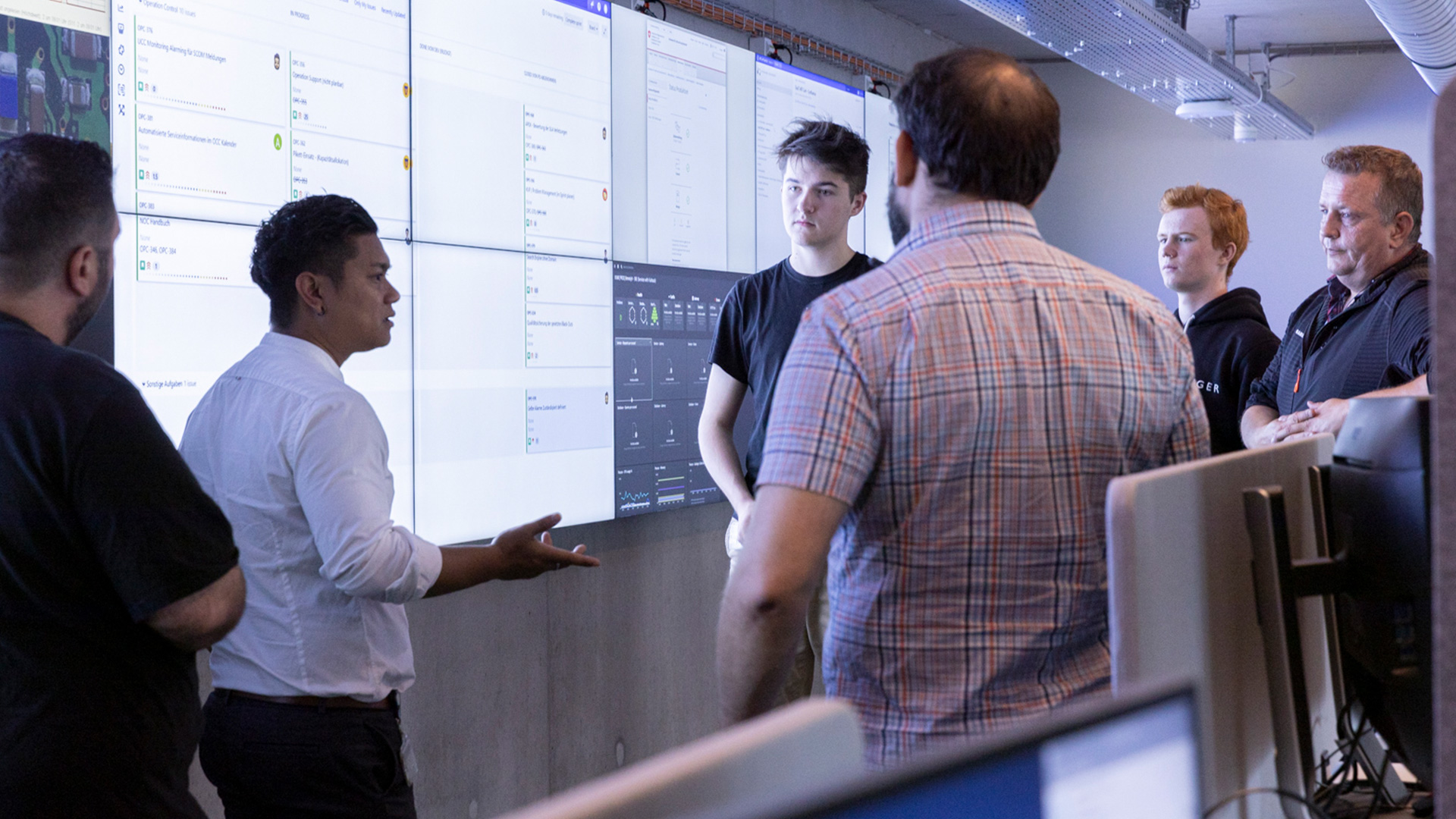 Eine Person erklärt mehreren Personen etwas vor einer Wand mit mehreren Bildschirmen.