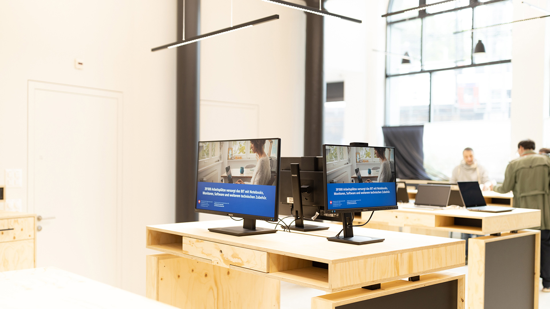 BIT-Store: Im Vordergrund stehen drei Bildschirme auf einem Tisch, dahinter steht ein weiterer Tisch mit drei Laptops. Im Hintergrund stehen beim Empfang drei Personen.