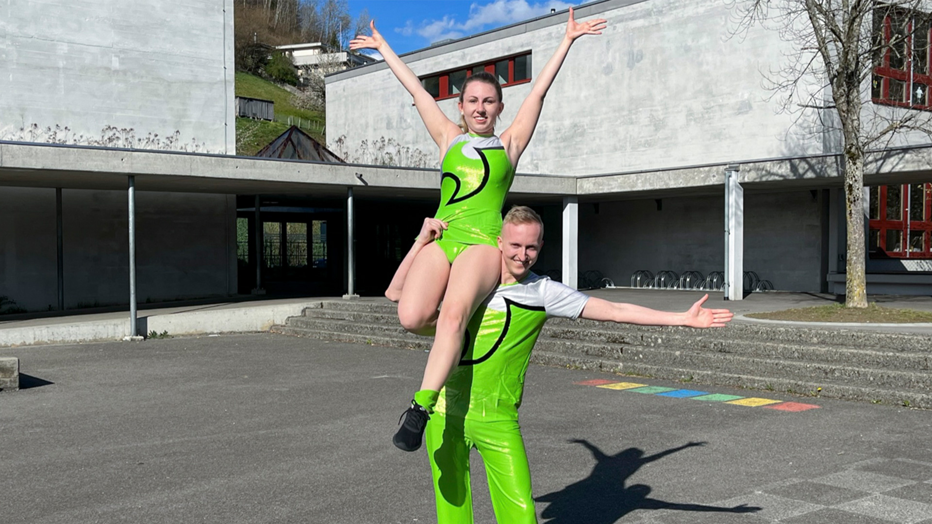 Tanzfigur: Mann hebt Frau mit angewinkeltem rechten Bein hoch. Sie stützt sich auf seiner rechten Schulter ab und streckt beide Arme aus.