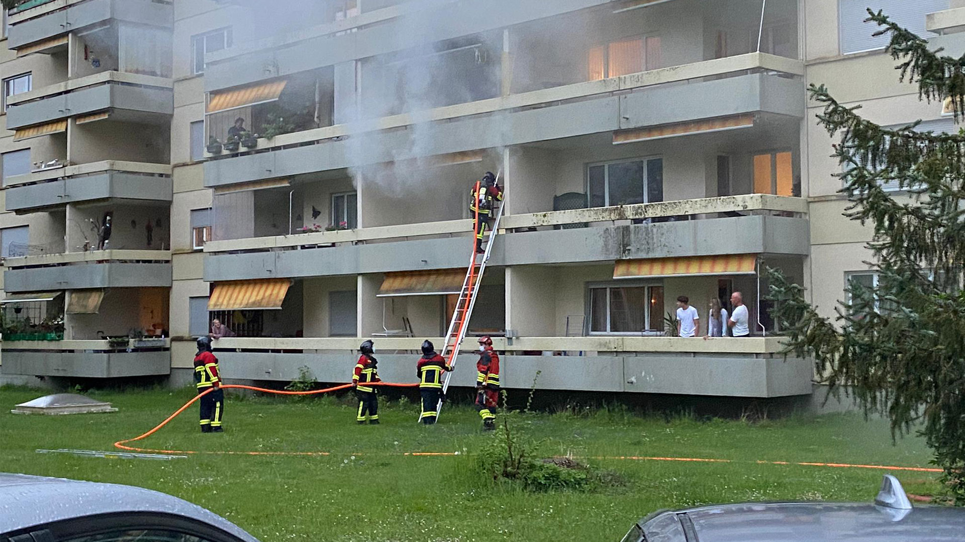 Fünf Feuerwehrleute löschen mit einem Strahlrohr ein Feuer in einem Wohnblock. Nachbarn stehen auf den Balkonen und sehen zu.