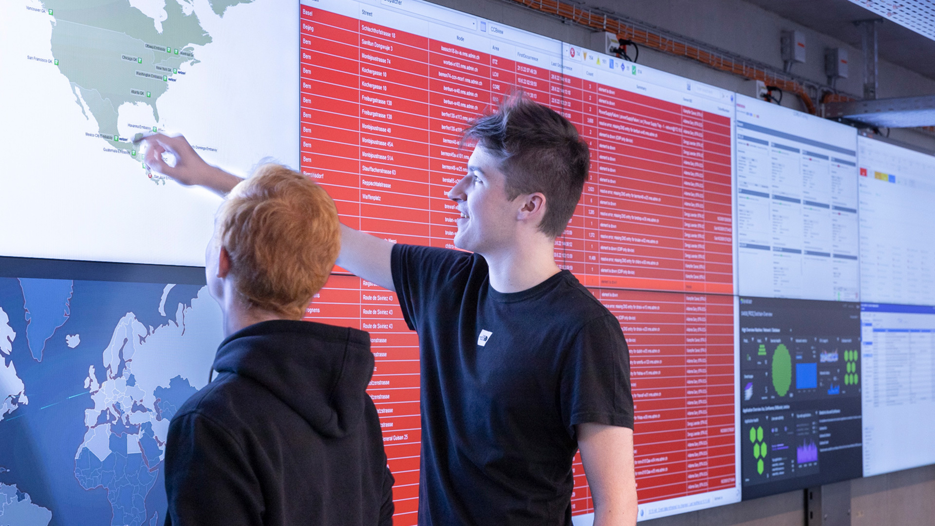 Une personne pointe à l’attention d’une autre l’un des écrans de la longue rangée de moniteurs fixés au mur.
