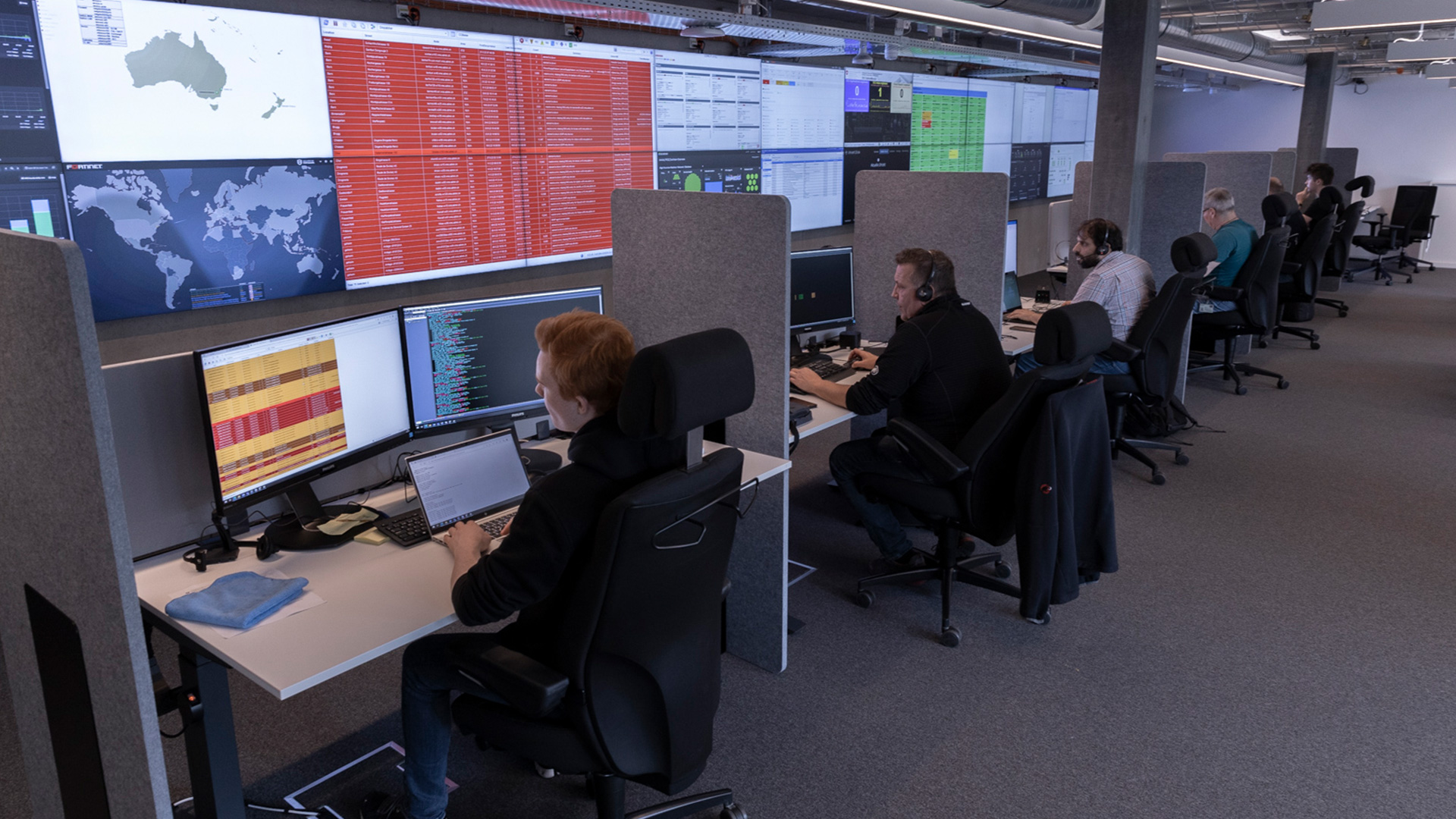 Centre de contrôle des opérations accueillant six personnes assises côte à côte devant leurs écrans personnels ainsi qu’une longue rangée d’écrans fixés au mur.
