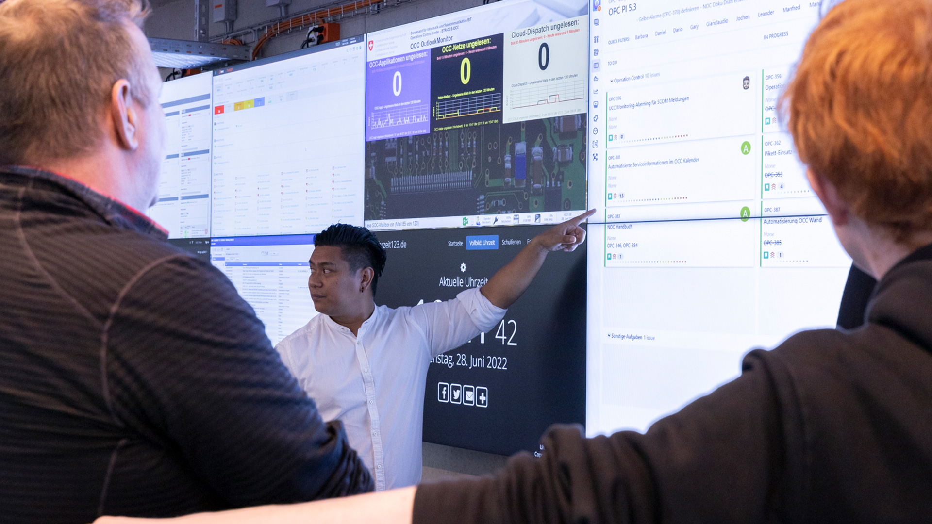 Une personne désigne l’un des écrans fixés au mur. Au premier plan, on distingue deux personnes assistant à sa présentation.